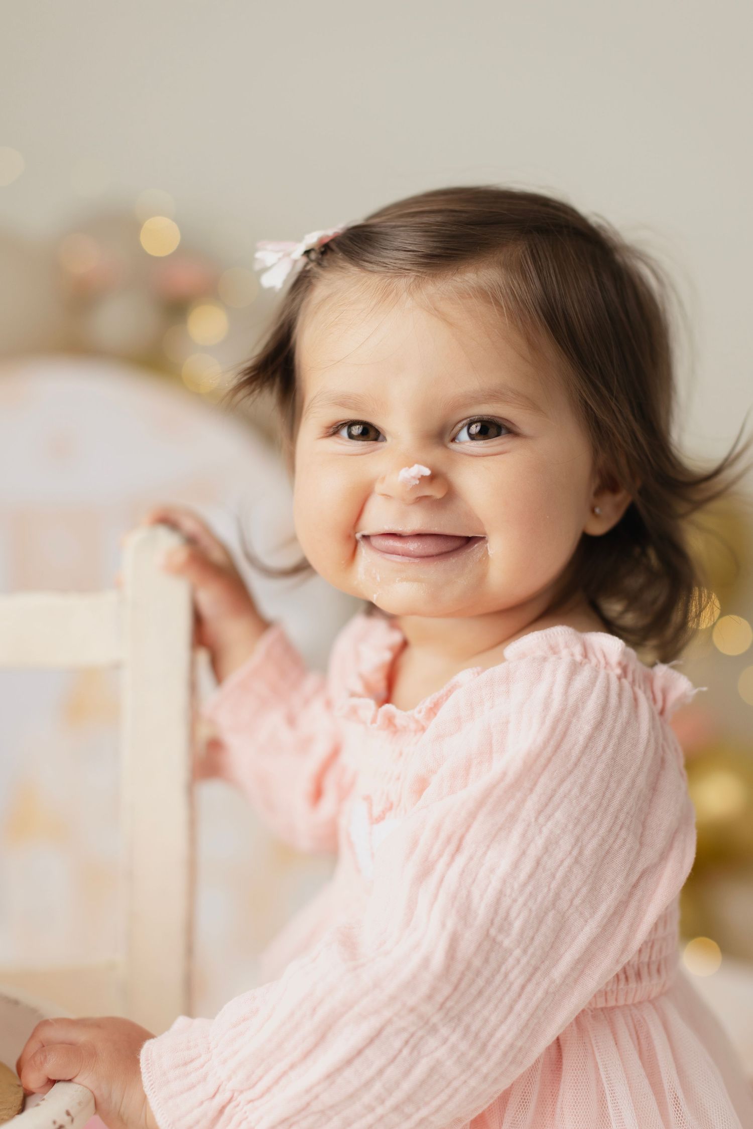 Smiling toddler girl with dark hair and white bow wearing pink dress with frosting on her nose. Elizabeth Lang Photography Surrey