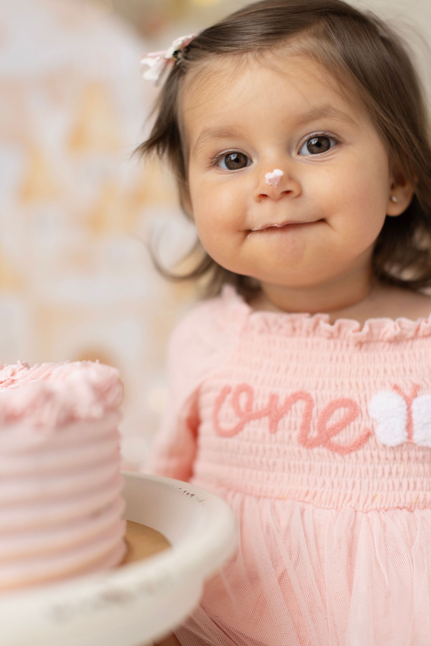 Smiling baby girl in pink smocked dress with cake on her first birthday. Elizabeth Lang Photography Surrey