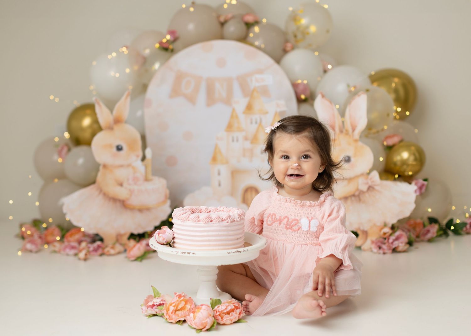 Baby girl in pink dress smiles beside a pink birthday cake, surrounded by bunny decorations and balloons. Elizabeth Lang Photography Surrey