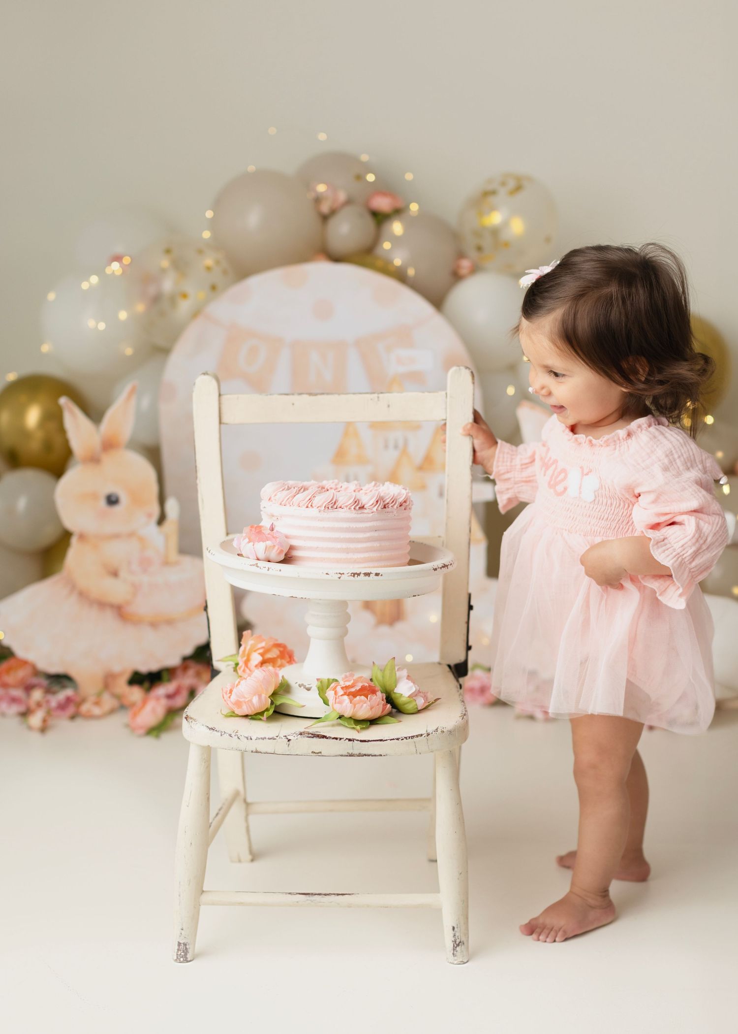 Toddler girl in pink dress standing beside white chair with pink birthday cake at a bunny-themed first birthday cake smash. Elizabeth Lang