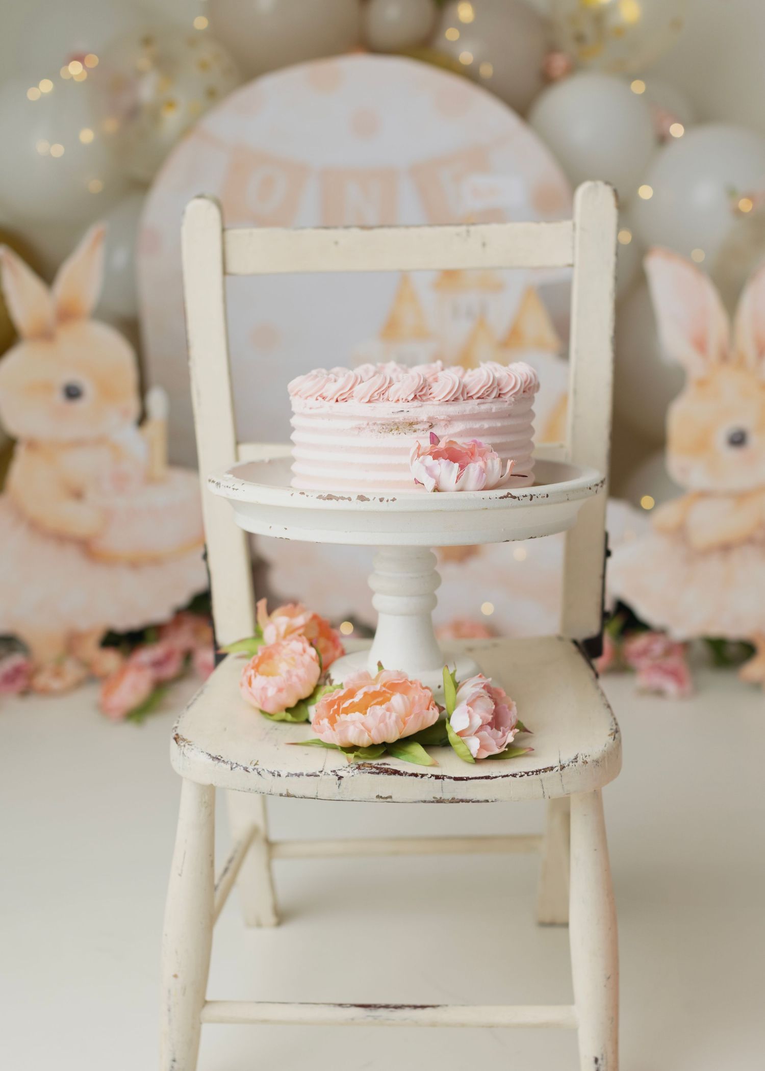 Pink cake on white pedestal chair with bunny decor and floral accents for a first birthday celebration. Elizabeth Lang Photography Surrey