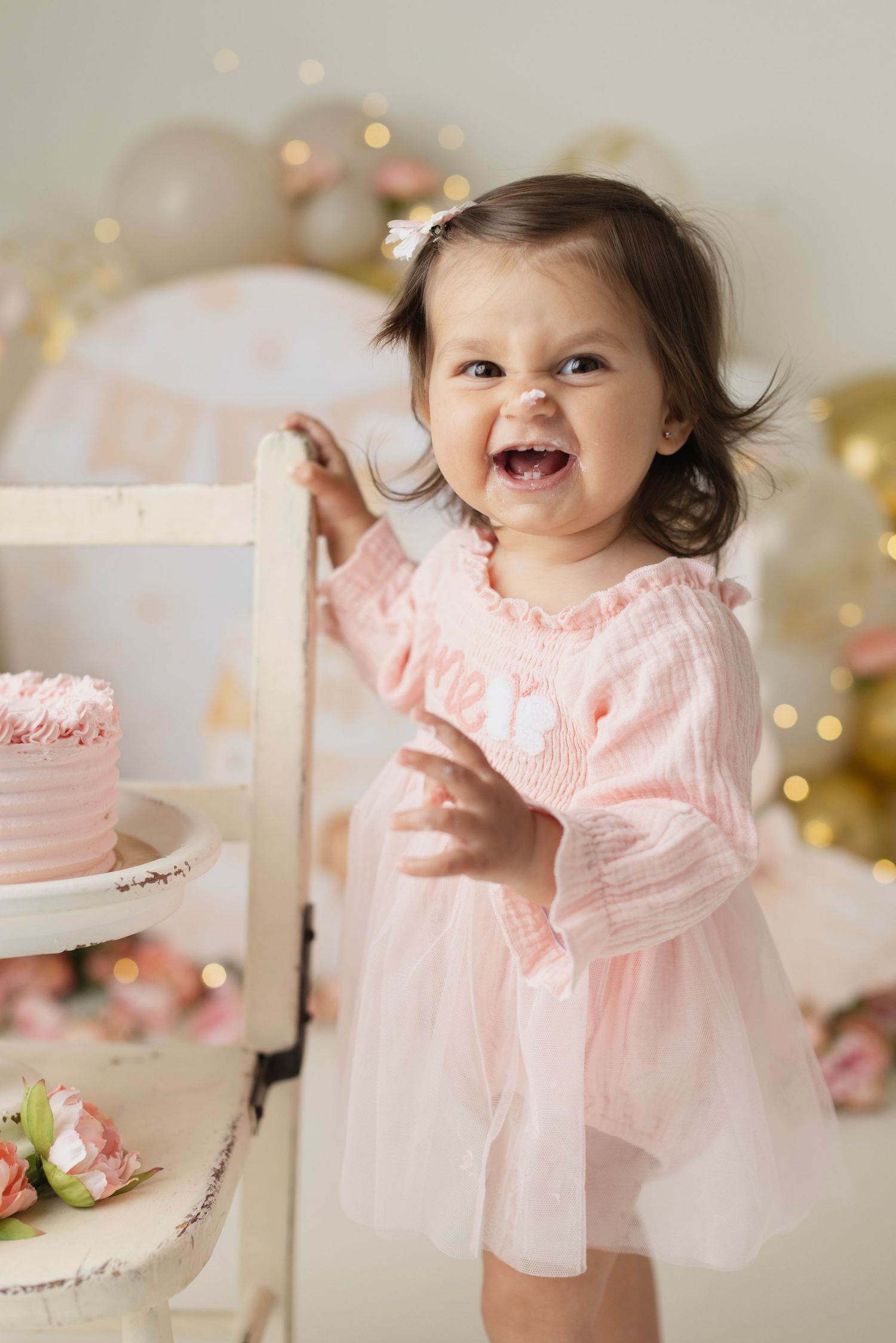 Smiling baby girl in pink dress celebrates first birthday beside a pink cake on a white stand. Elizabeth Lang Photography Surrey