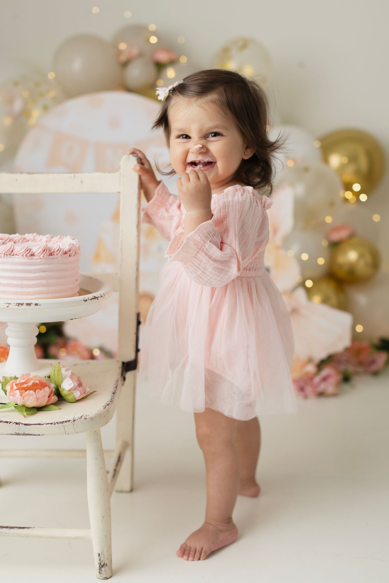 Happy toddler in pink tutu dress standing by birthday cake at party. Elizabeth Lang Photography Surrey