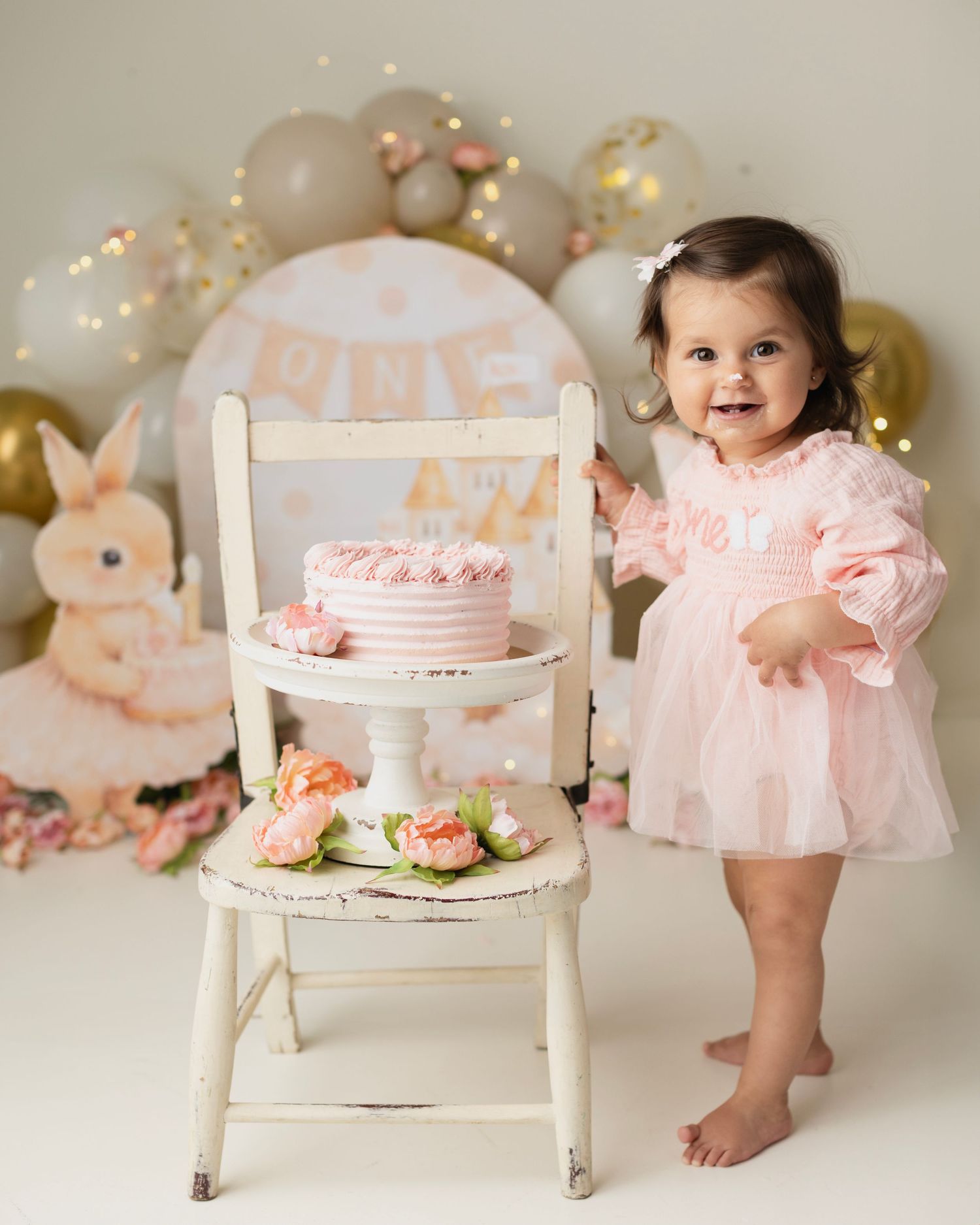 Smiling toddler girl in pink dress standing next to white chair with pink cake at a bunny-themed first birthday celebration. Elizabeth Lang