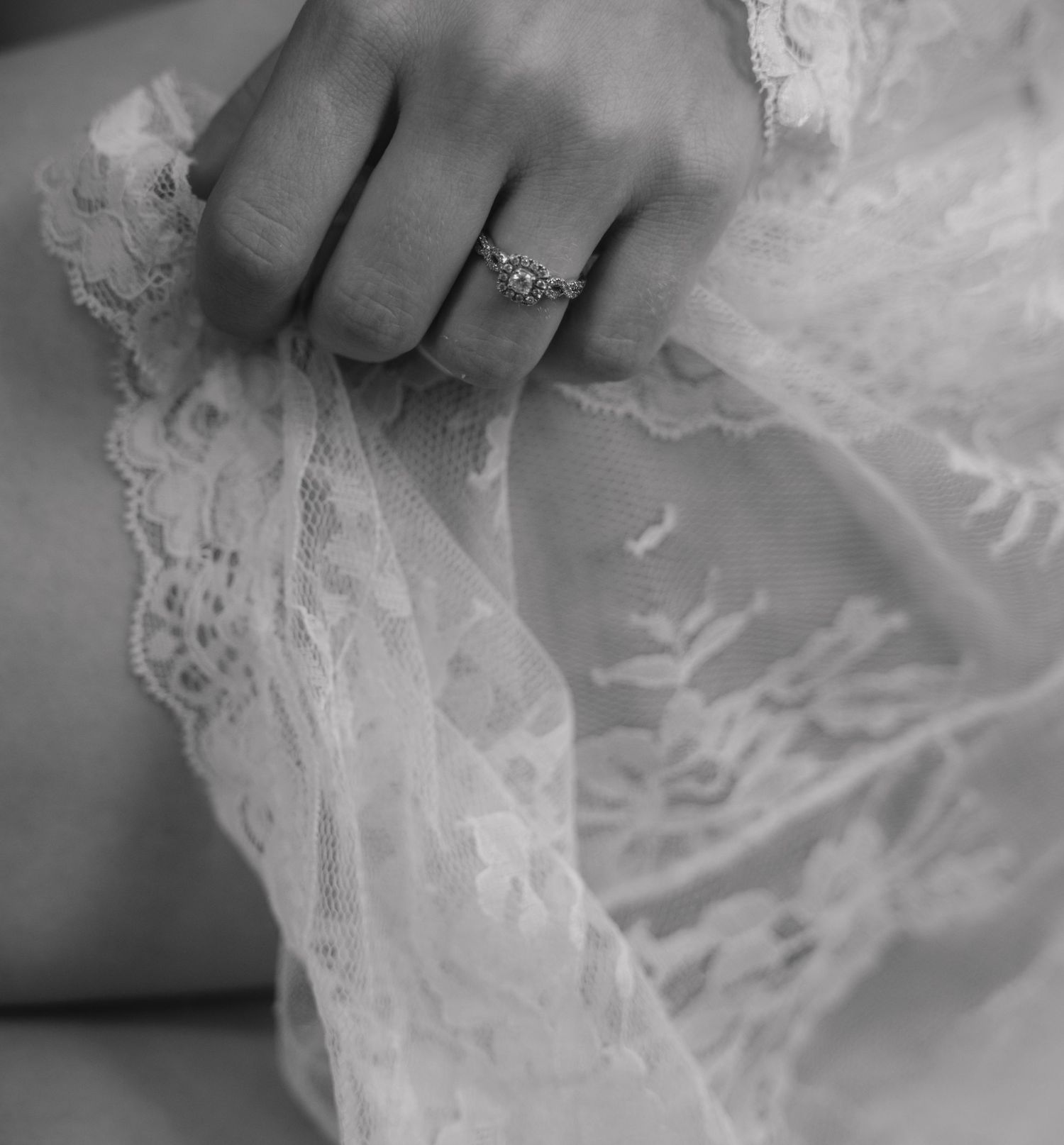 Black and white photo of a hand wearing a diamond ring, delicately holding white lace fabric.