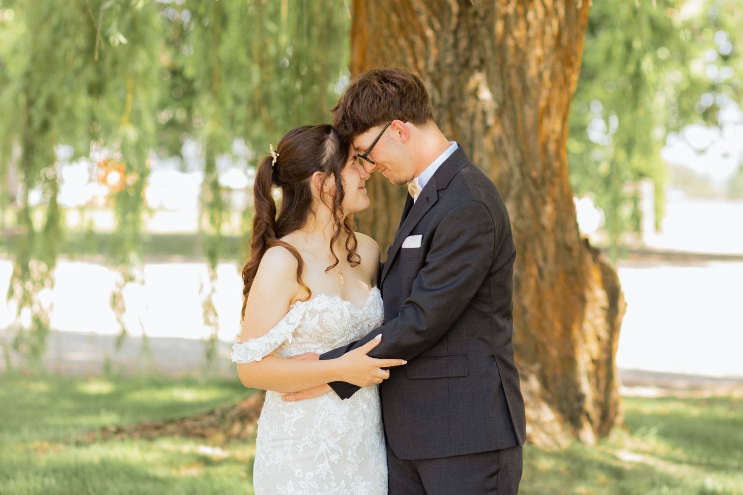 A bride and groom share a romantic moment under a weeping willow tree outdoors on their wedding day.