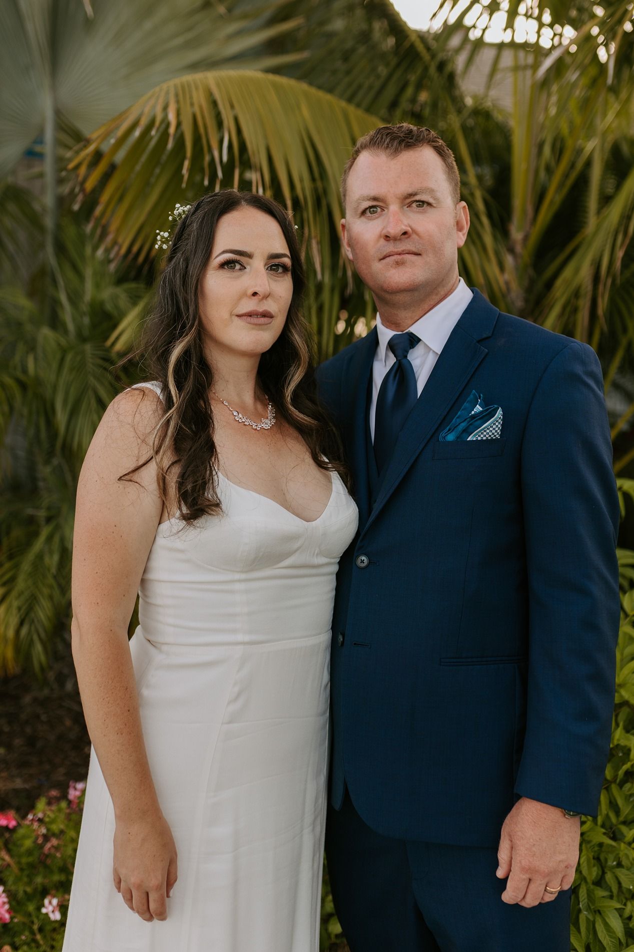Bride in white dress and groom in navy suit pose together outdoors with tropical palm trees in background at Tower Beach Club.