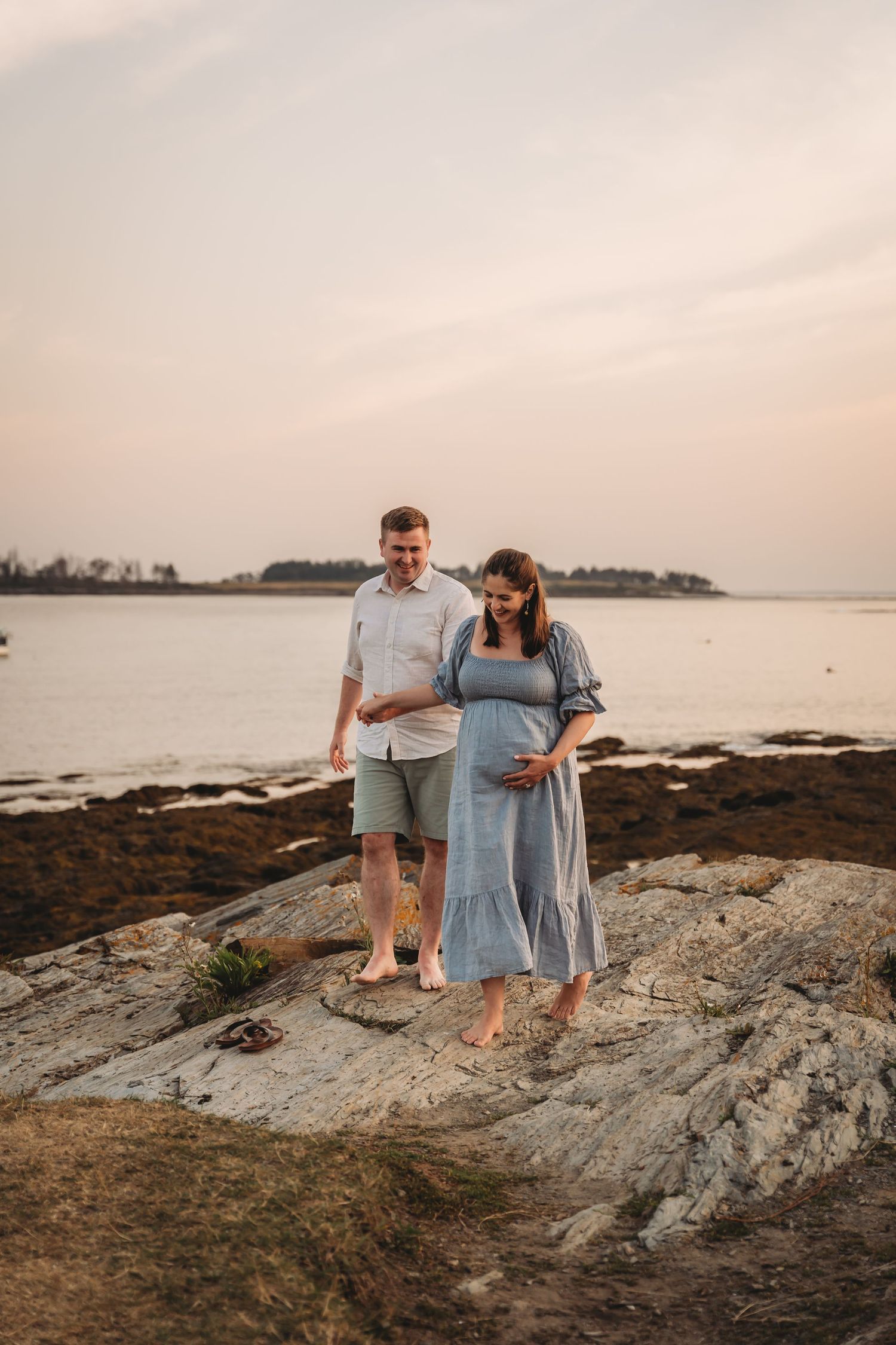 Pregnant woman in white dress standing on rocky beach at golden sunset, gently cradling her belly.