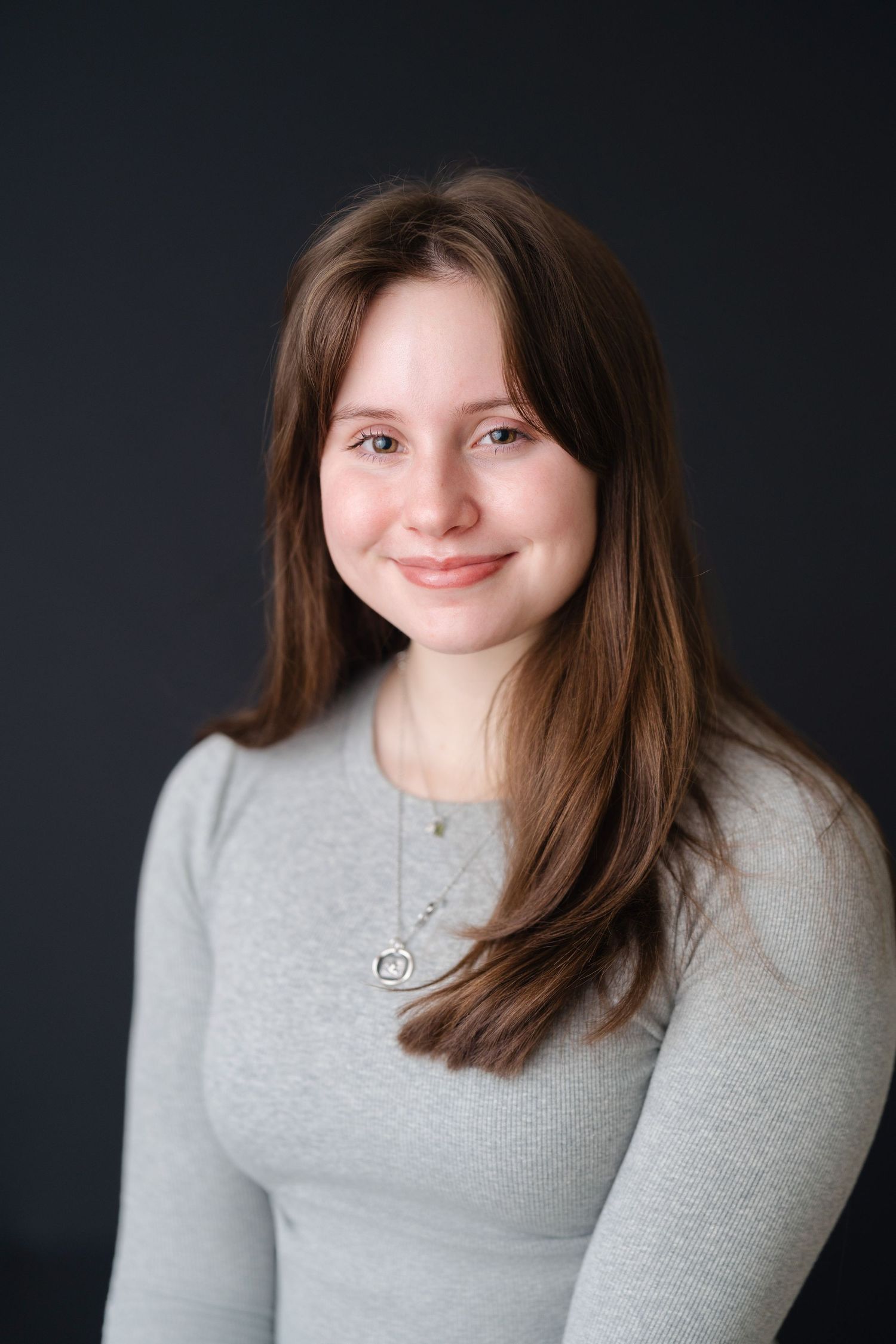 Young woman with long brown hair smiling, wearing a white knit cardigan and silver necklace against dark background.