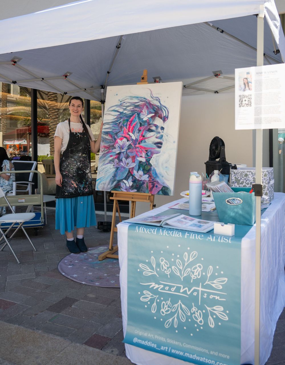 Artist stands beside colorful portrait painting at outdoor art booth with display table and supplies.