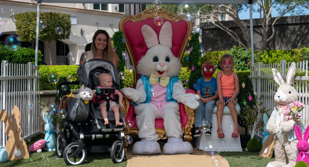 Mother with children including stroller pose with Easter Bunny on decorated throne at spring event.