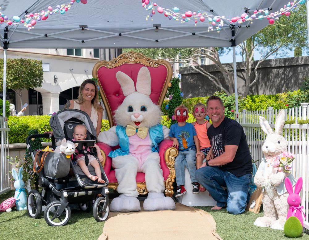 Family with children poses with Easter Bunny on throne at outdoor Easter event with festive decorations.