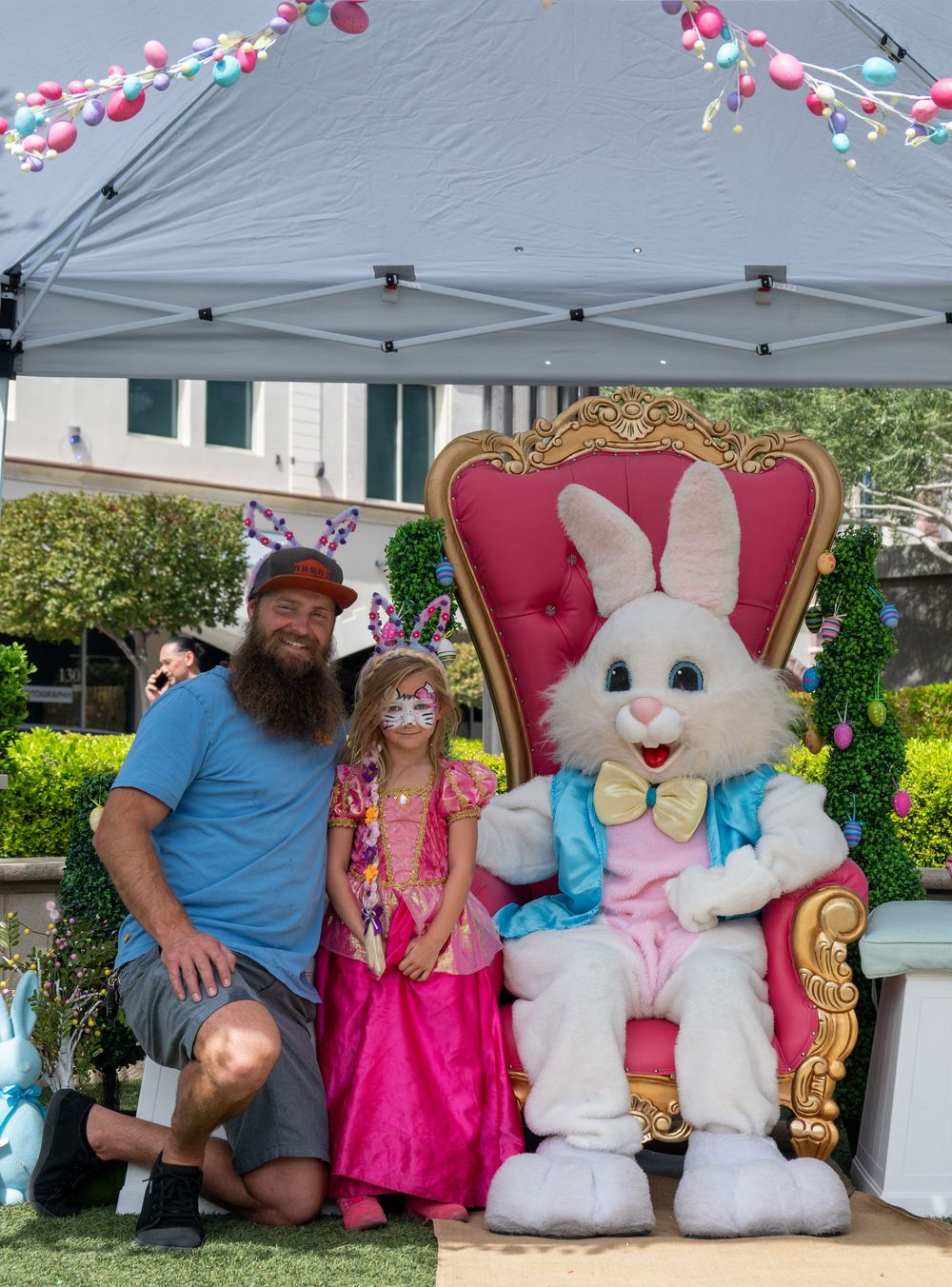 Father and daughter in pink dress pose with Easter Bunny on decorative throne at outdoor event.