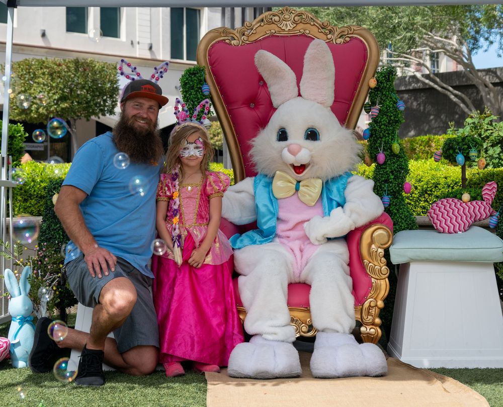Man kneeling with young girl in pink dress beside Easter Bunny on gold and pink throne outdoors.