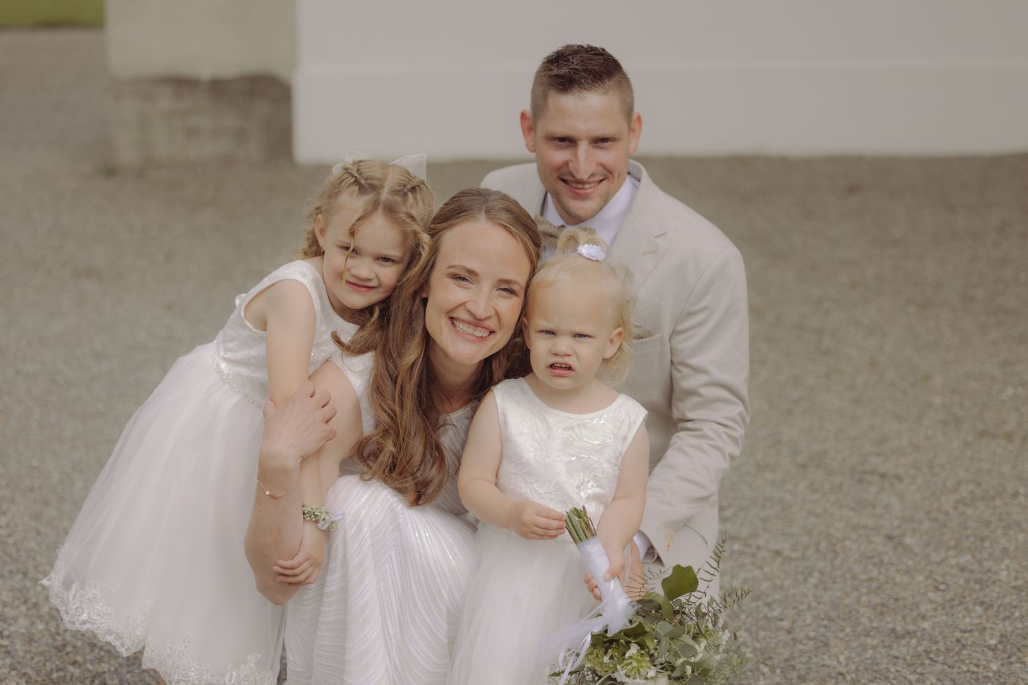 Happy family of four posing together at a wedding, with two young girls in white dresses.