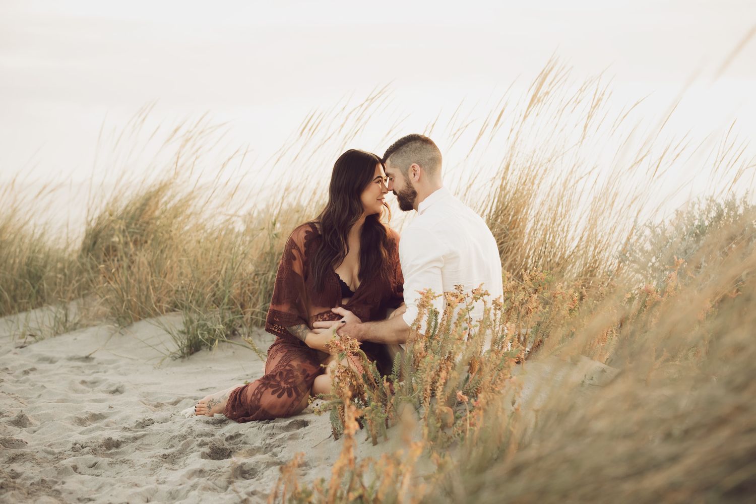 Expecting couple sitting together in sandy beach dunes with tall grass at soft golden hour sunset.