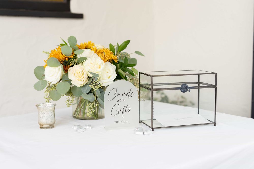 Wedding cards and gifts table with glass box, white sign, and yellow floral arrangement.