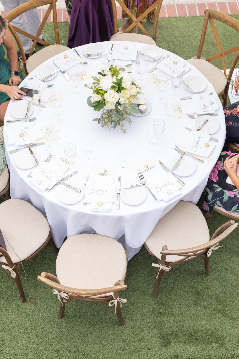 Elegant outdoor wedding reception table with white linen, floral centerpiece, and place settings on green grass.