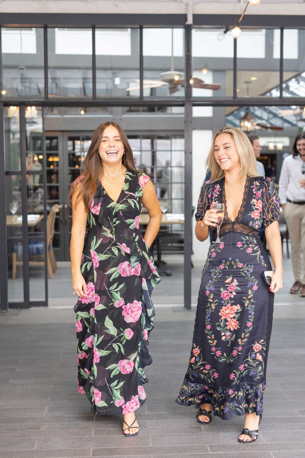 Two women in black floral maxi dresses laugh and walk together at an outdoor event.