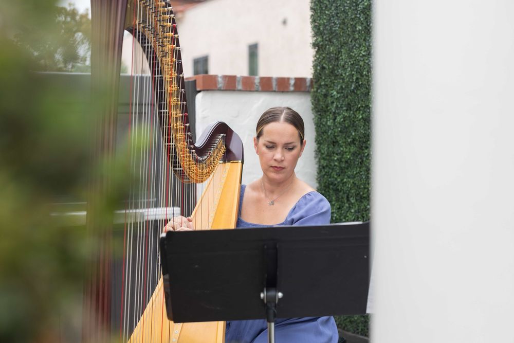 Female harpist in blue dress reads sheet music while playing harp outdoors.
