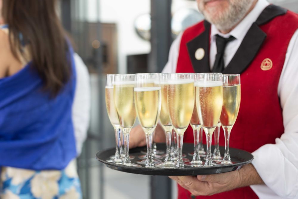 Waiter in red vest serves champagne flutes on a silver tray at a formal event.