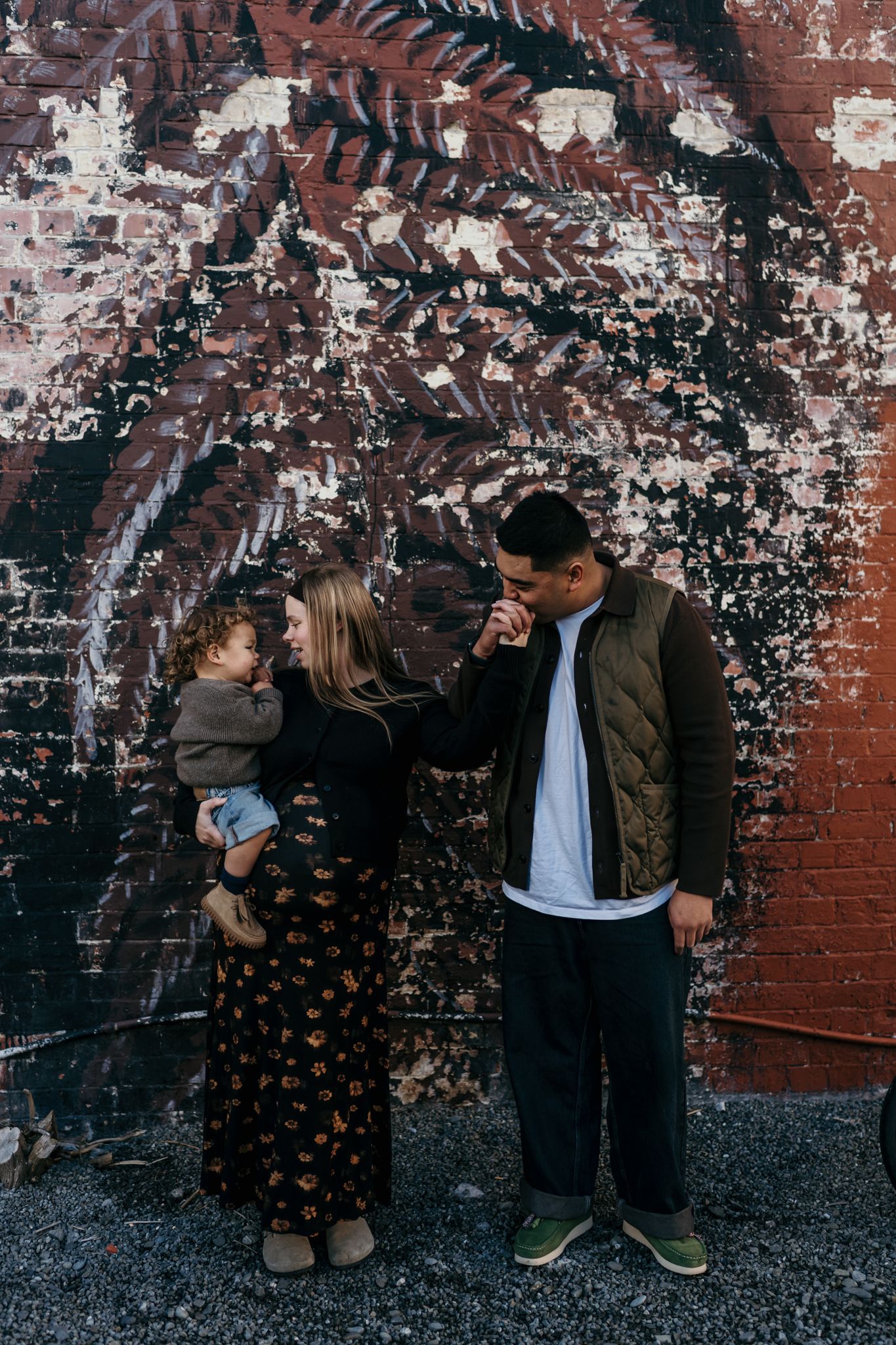 A young family poses in front of a weathered brick wall mural, woman holding toddler beside man in olive jacket.