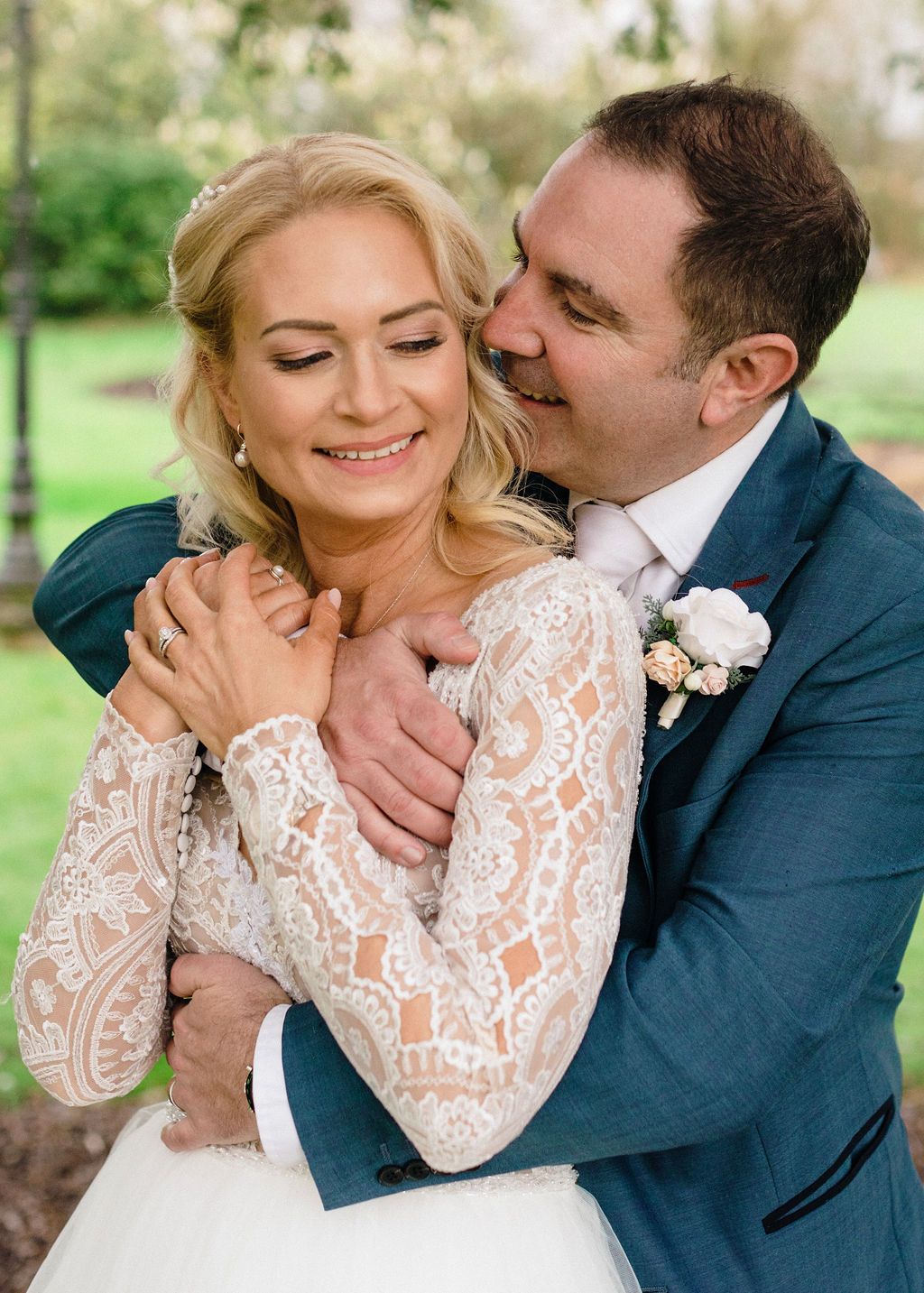 A joyful bride and groom embrace outdoors, she in lace wedding gown, he in teal suit with boutonniere.