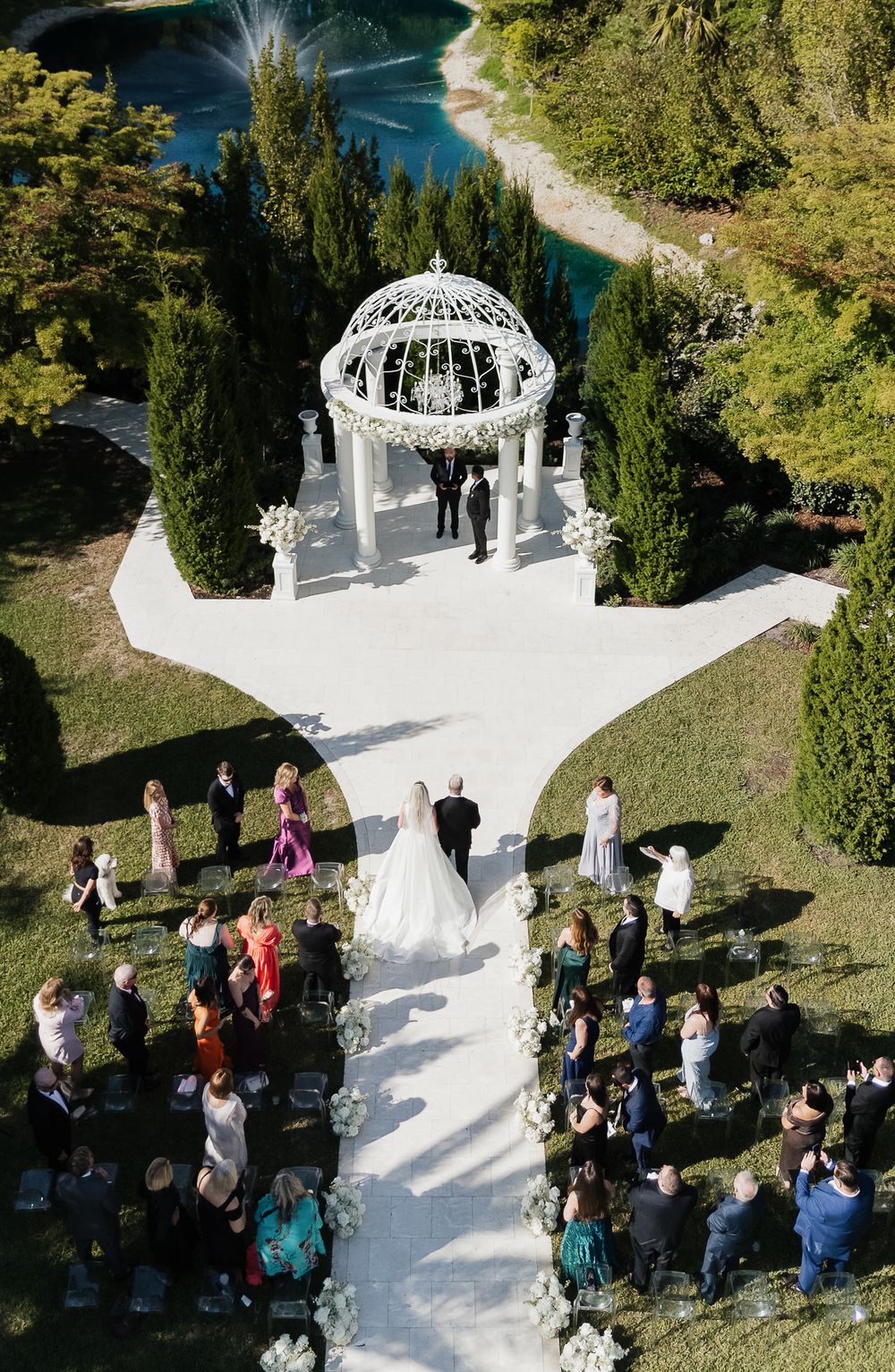 Aerial view of elegant outdoor wedding ceremony at white gazebo, bride and groom walking aisle with guests watching.