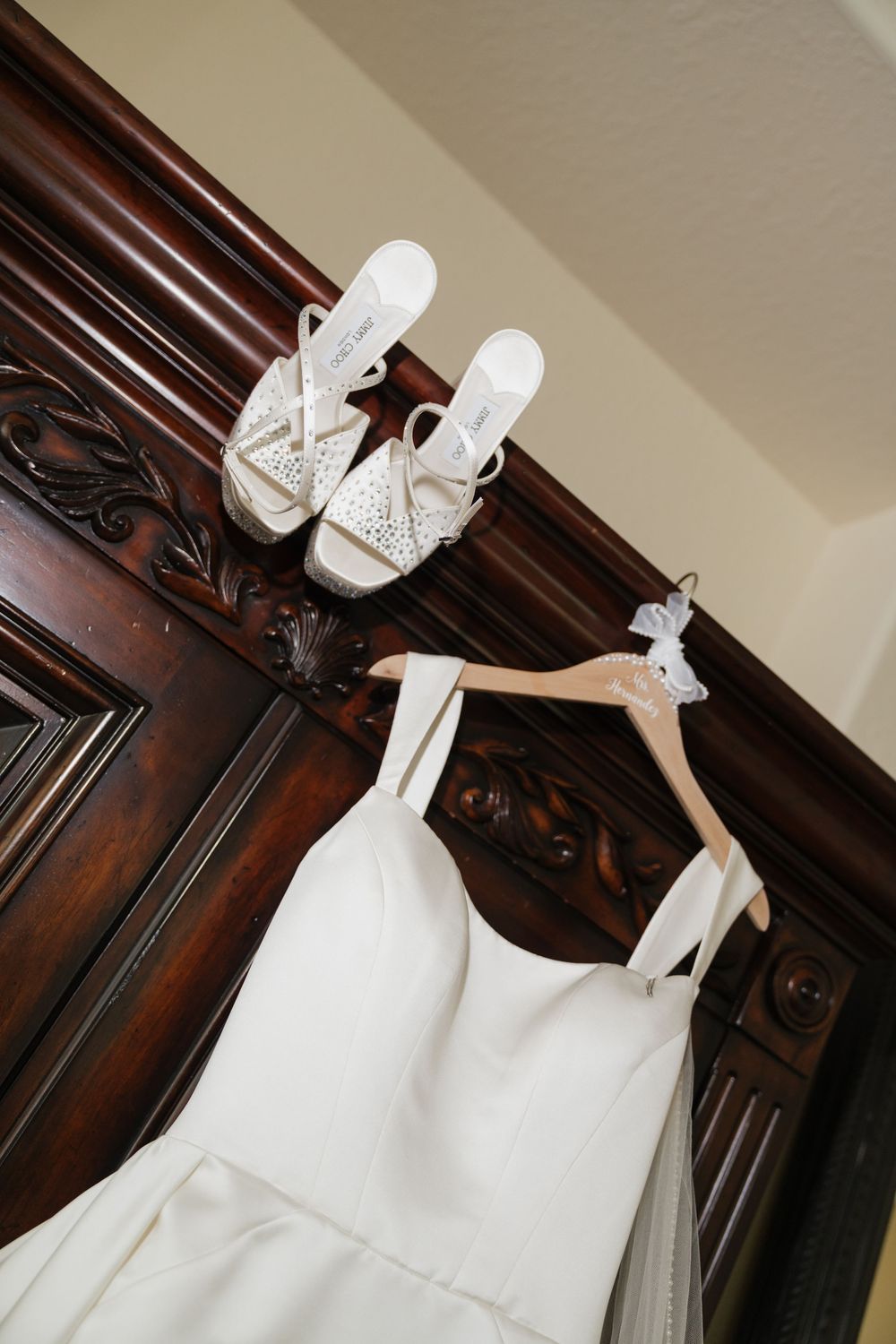 White wedding dress hanging on wooden armoire with matching rhinestone heeled sandals displayed above.
