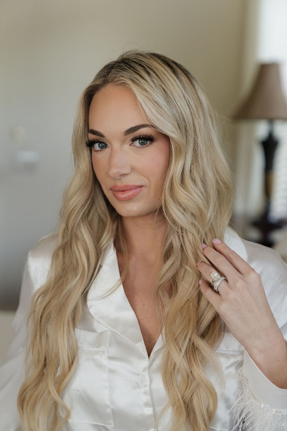 Blonde woman with long wavy hair wearing a white satin robe, showcasing a diamond ring while posing indoors.
