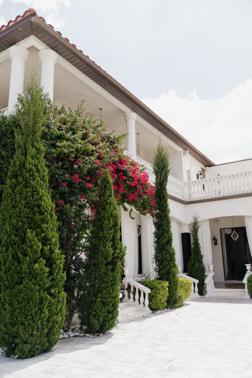 Elegant white villa with red-tiled roof, lush cypress trees, and vibrant bougainvillea climbing the columns in a manicured courtyard.