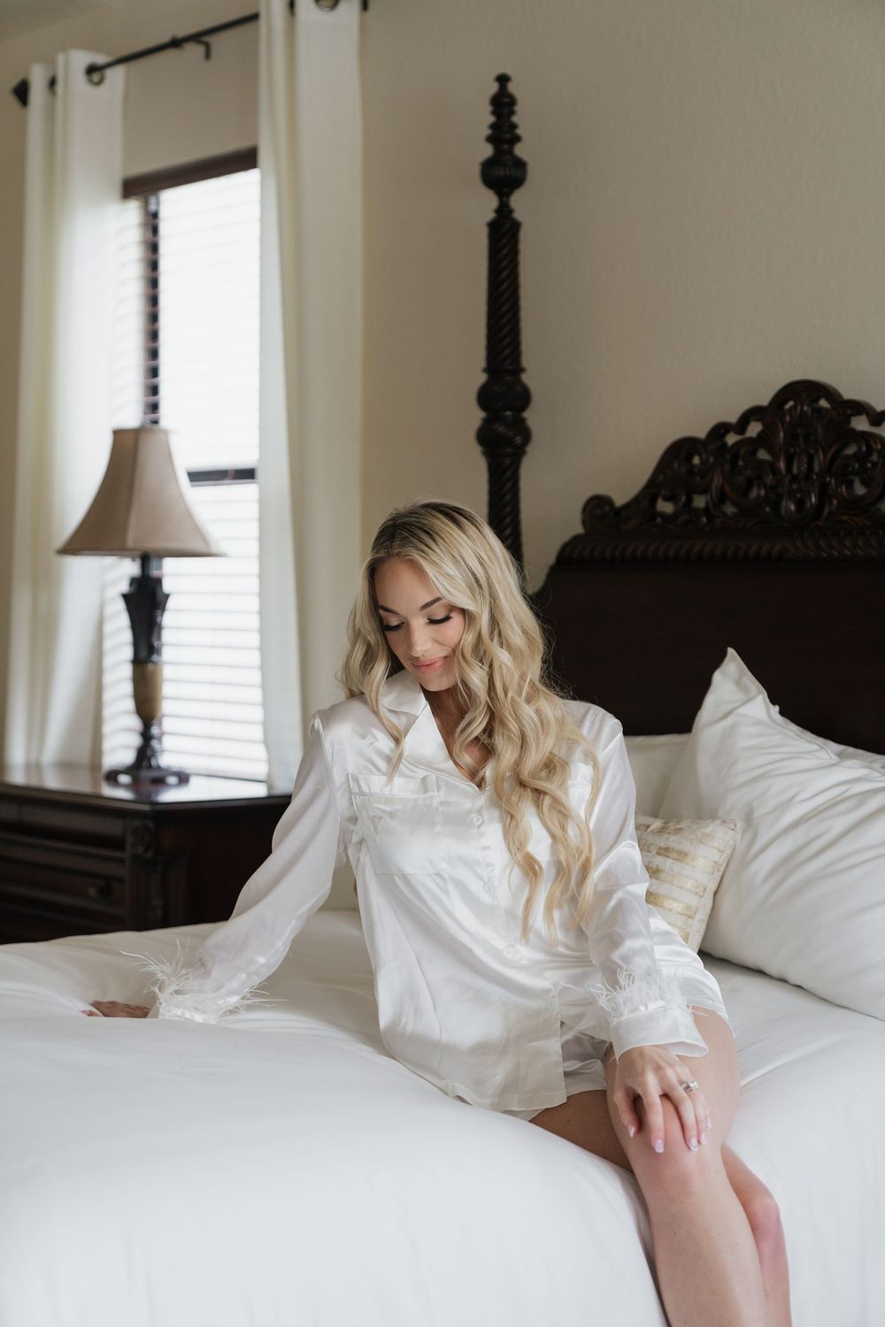 Blonde bride in white satin robe sitting on elegant bed during wedding morning preparations.
