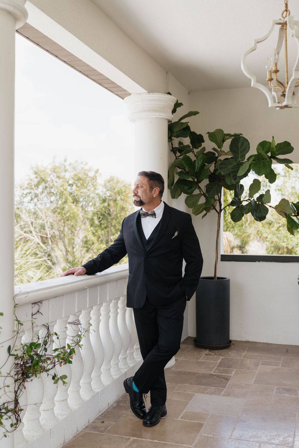 Dapper groom in black tuxedo with bow tie standing on elegant white balcony beside lush fiddle leaf fig tree.