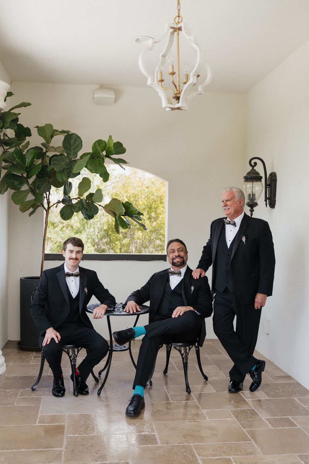Three men in black tuxedos posing indoors, two seated and one standing, before a wedding ceremony near a fiddle-leaf fig tree.