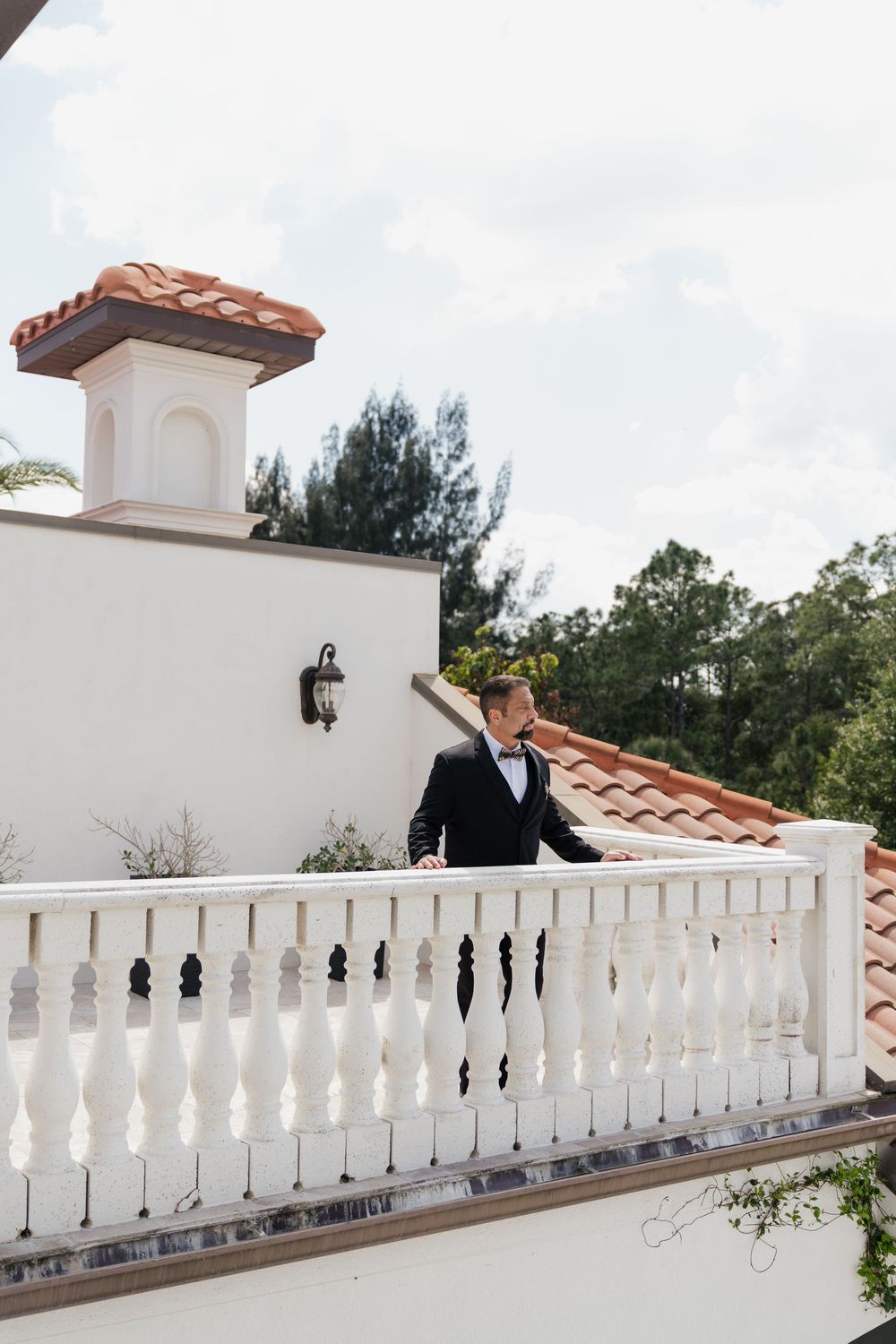 Dapper groom in black tuxedo standing on a white balustrade balcony of a Mediterranean-style villa with terracotta roof tiles.