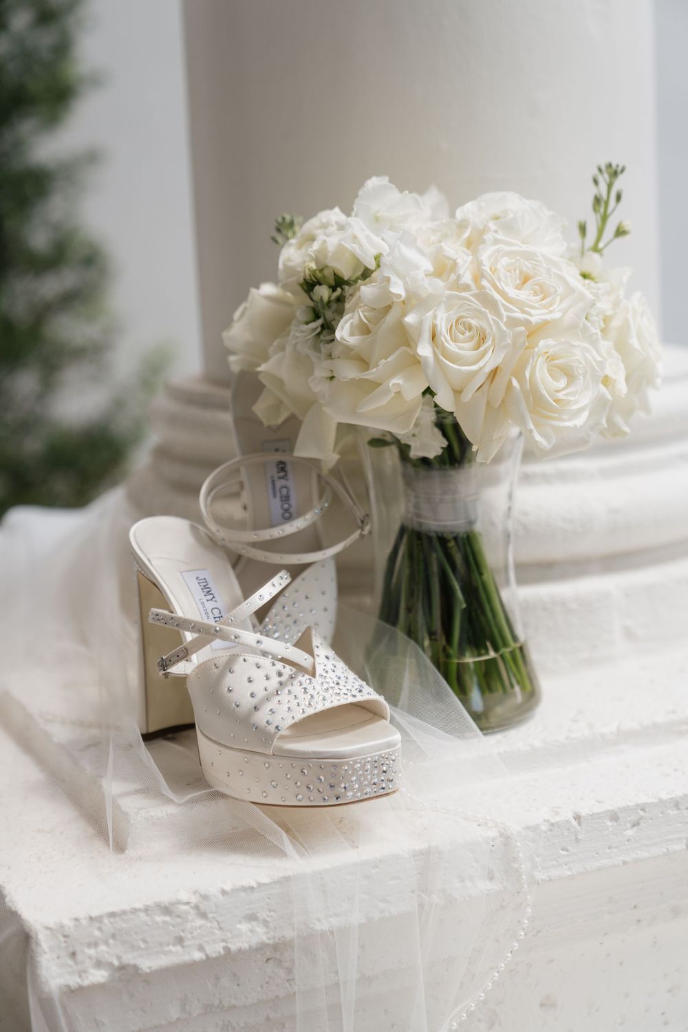 White Jimmy Choo platform wedding shoes with crystal embellishments beside a bouquet of white roses in a vase.
