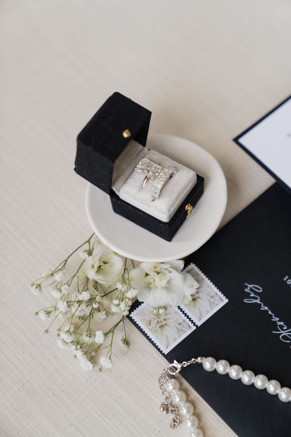 Elegant wedding ring in black velvet box on white dish, with baby's breath flowers, pearl necklace, and dark invitation.