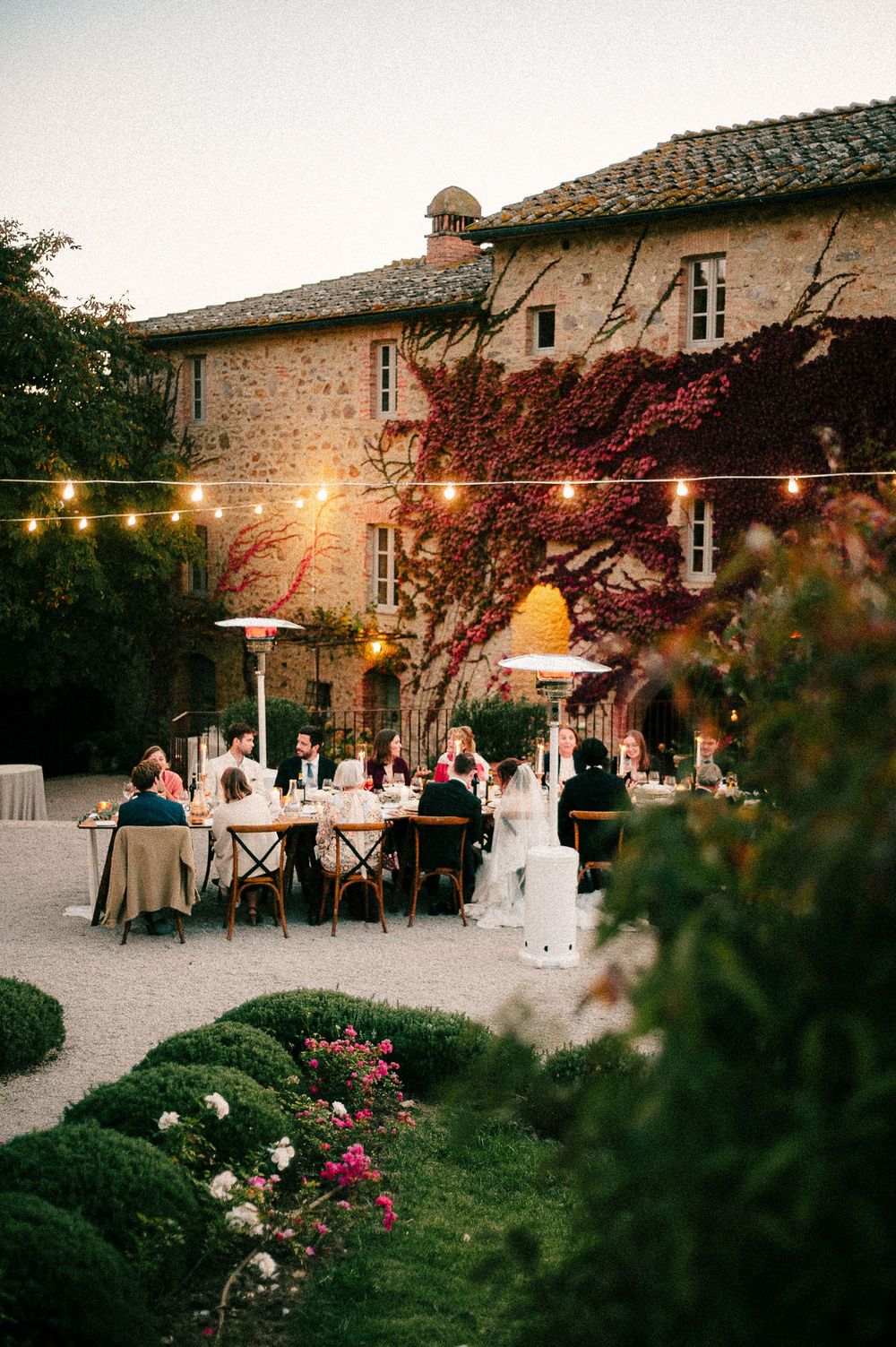 Elegant outdoor wedding reception at a rustic stone villa with string lights, lush greenery, and guests dining at dusk.