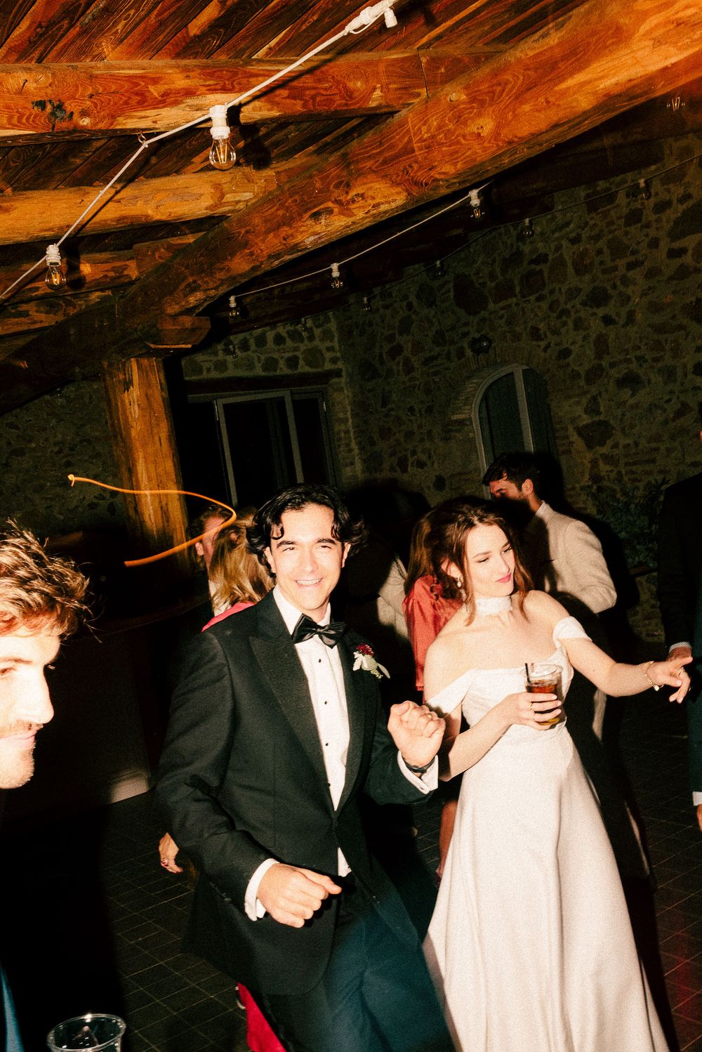 Bride and groom dancing joyfully at wedding reception in rustic stone venue with string lights overhead.