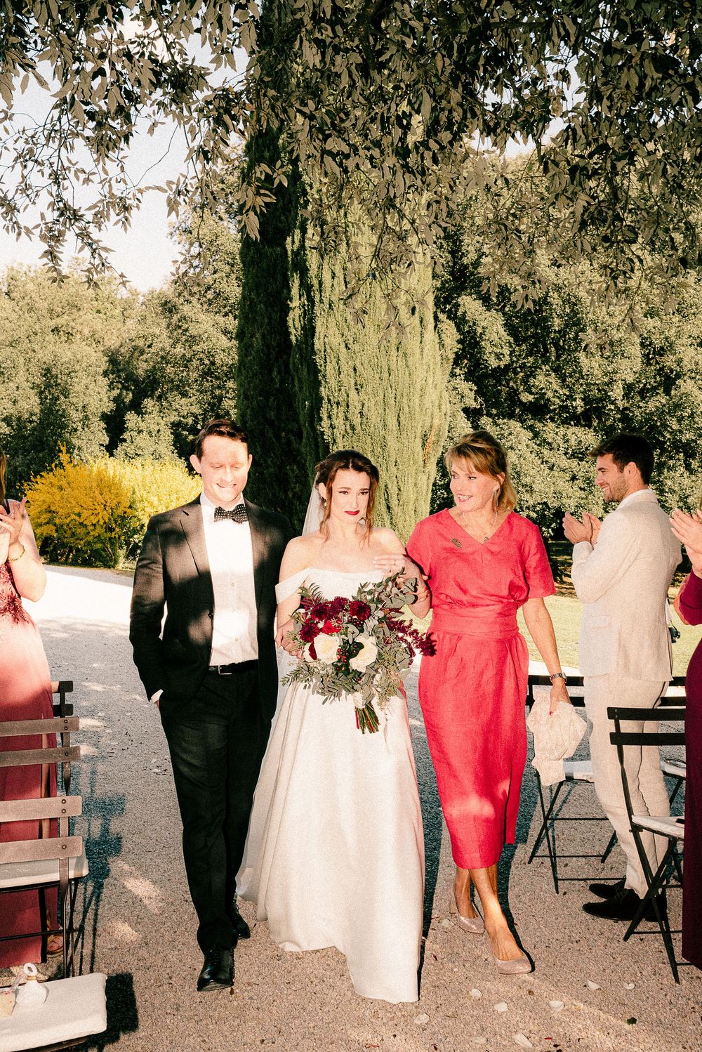 Bride and groom walk down the aisle with a woman in red dress at an outdoor wedding ceremony.
