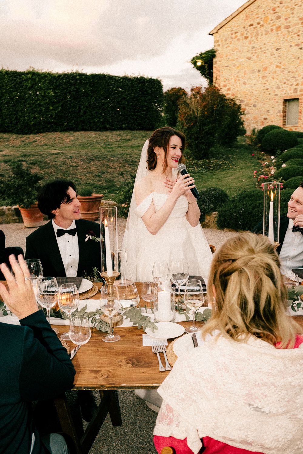 Bride in white gown gives speech at outdoor wedding reception table with guests in formal attire.