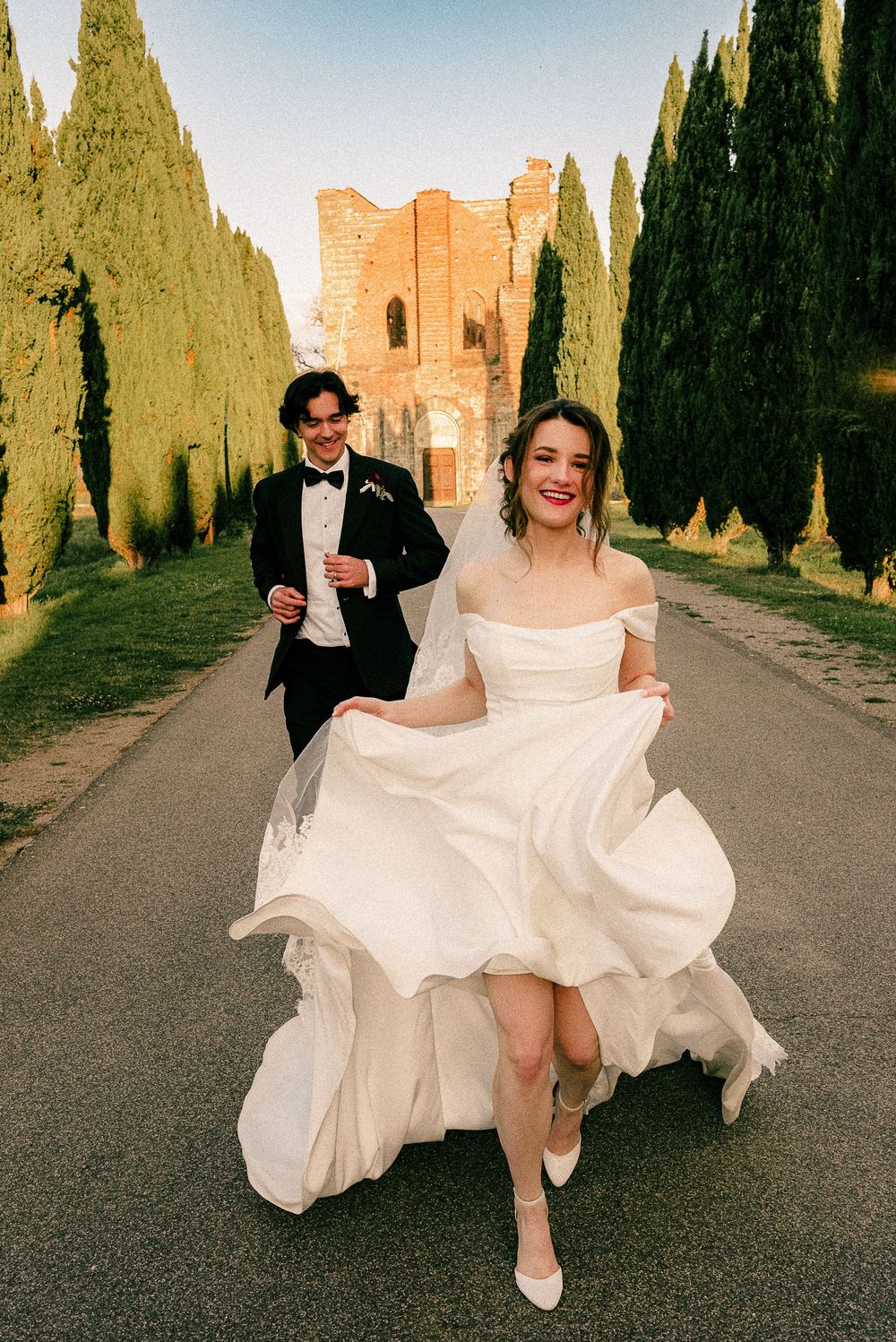 Bride in white off-shoulder gown lifts dress while running with groom in tuxedo along cypress-lined path in Tuscany.