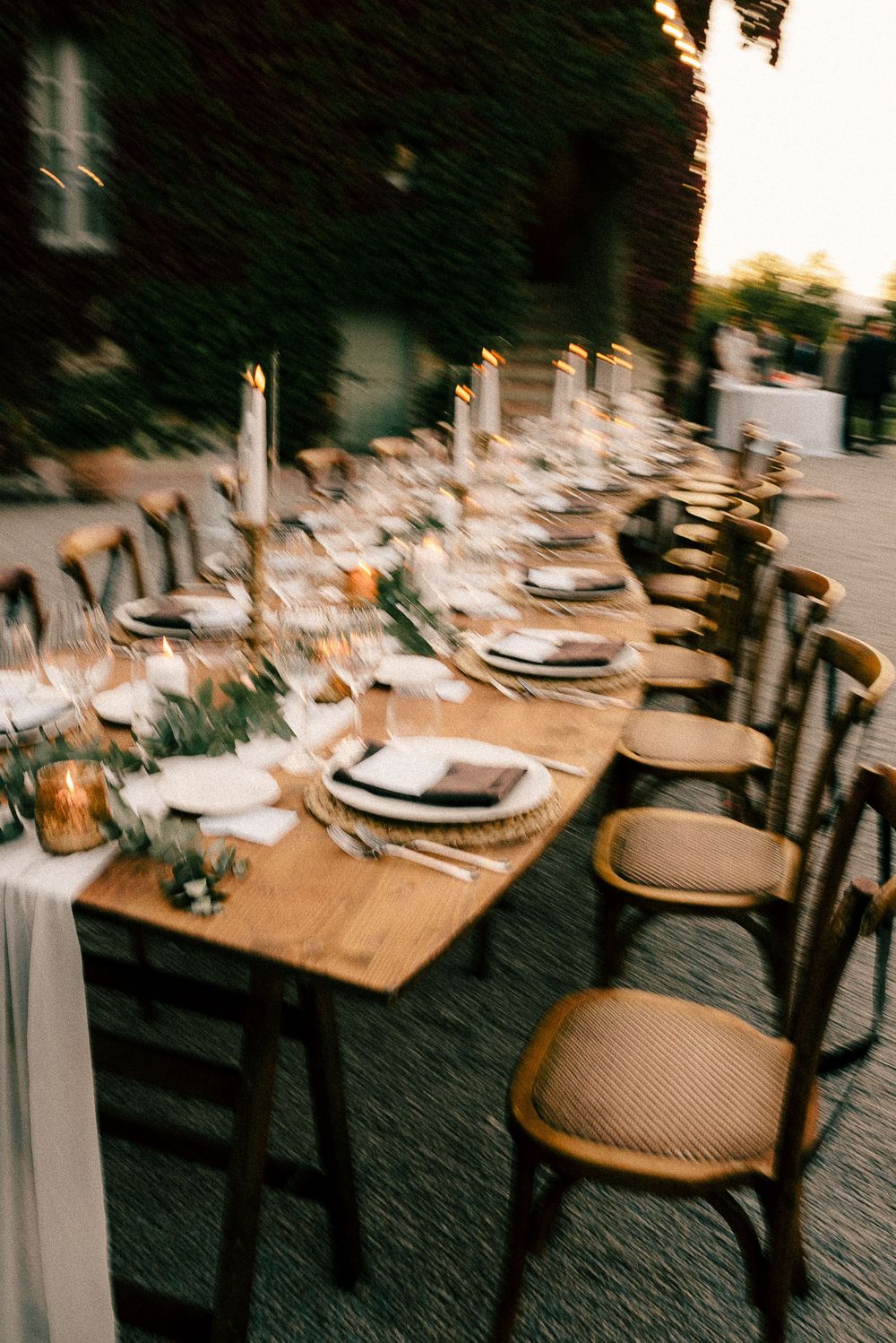 Elegant outdoor wedding reception with long wooden table, candles, greenery, and wicker chairs at golden hour.