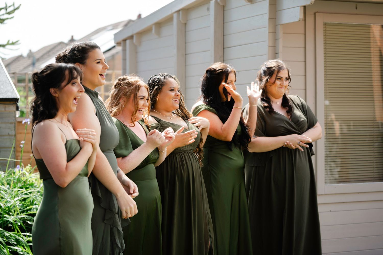 Six bridesmaids in sage and olive green dresses standing in a row, emotionally reacting during a wedding ceremony outdoor.