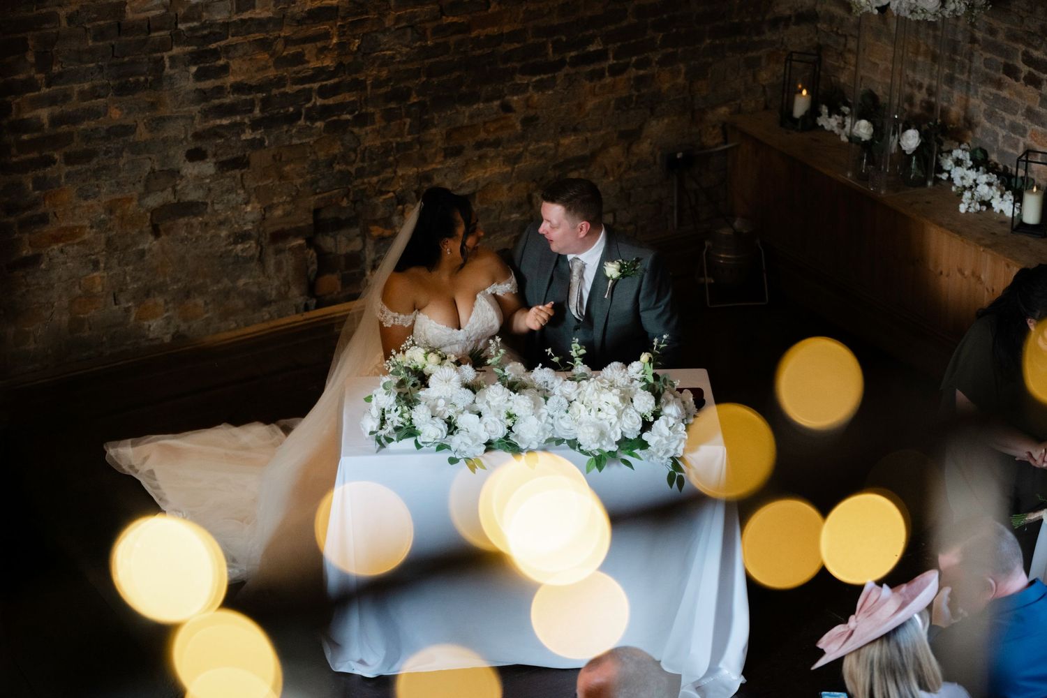Bride and groom share a romantic moment at their floral wedding table, surrounded by bokeh lights in a rustic brick venue.