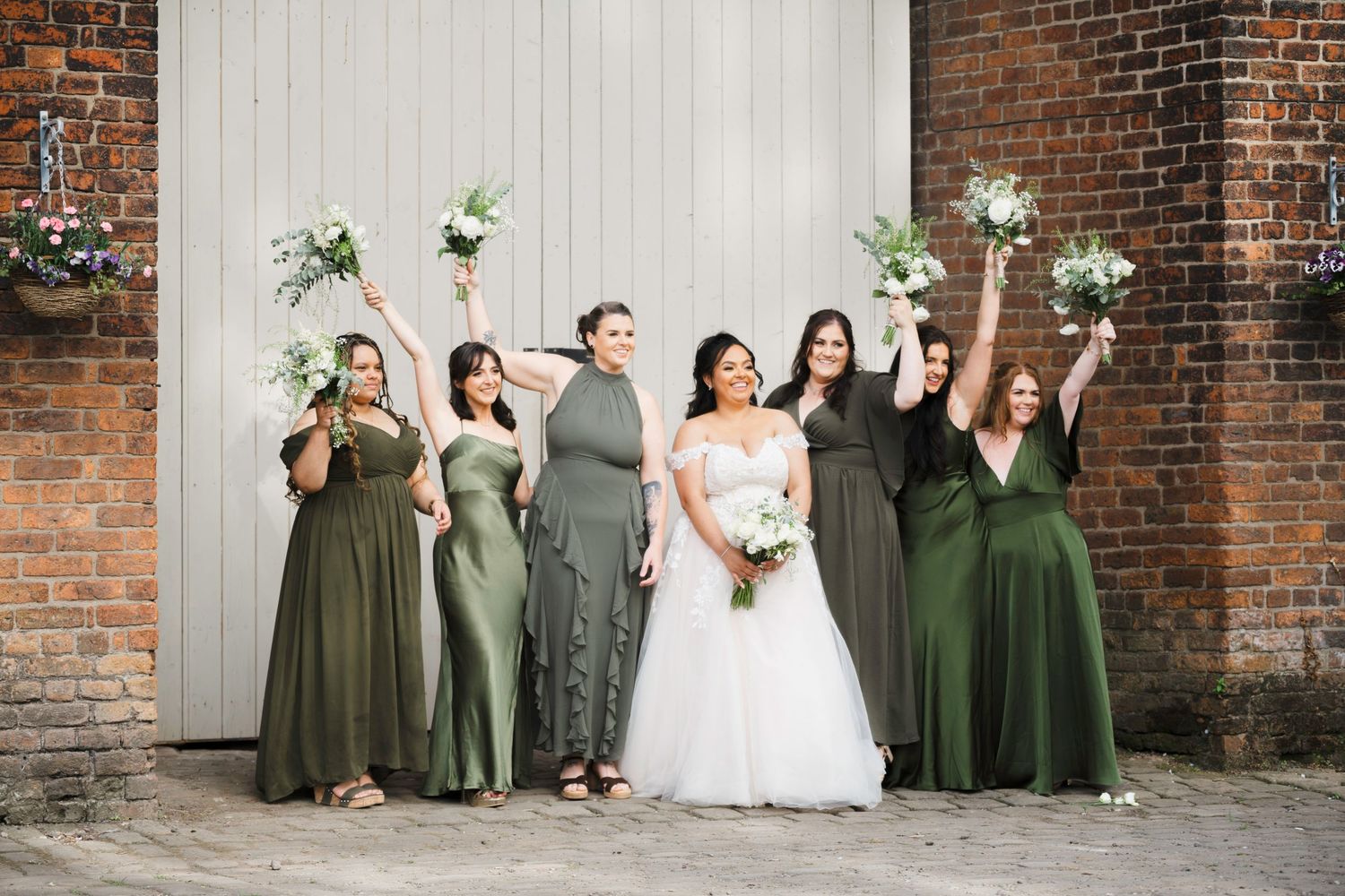 Bride in white gown with five bridesmaids in green dresses celebrating outdoors near brick wall.