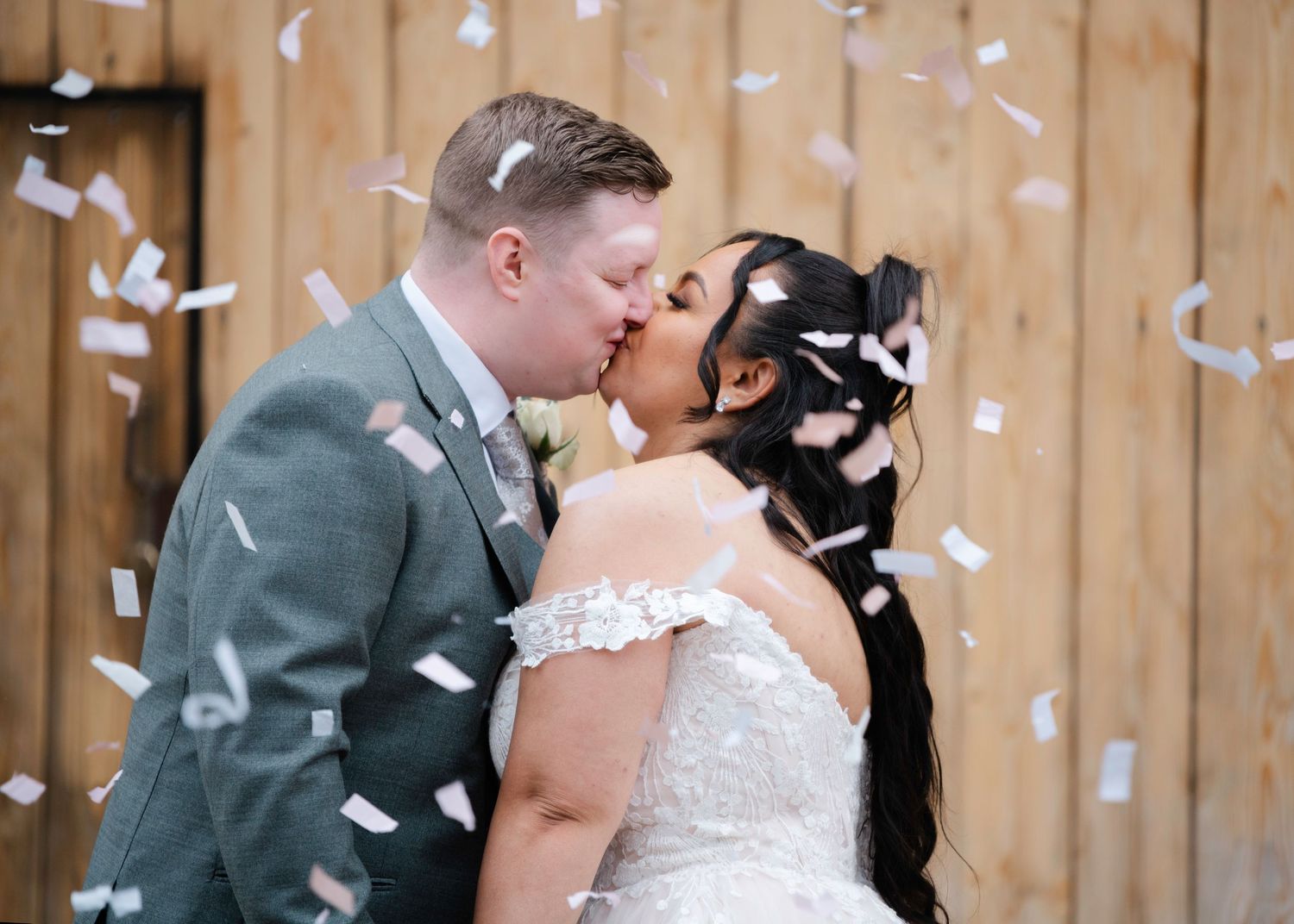 Newlywed couple sharing a kiss surrounded by falling confetti, bride in lace gown and groom in grey suit.