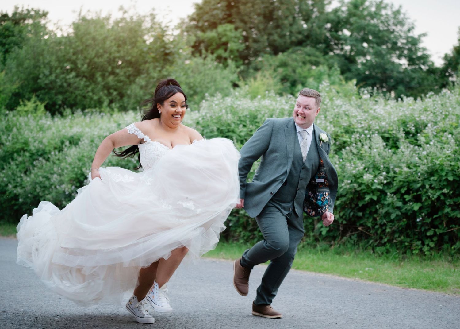Bride and groom laughing and running together outdoors on their wedding day, bride in white gown with sneakers.