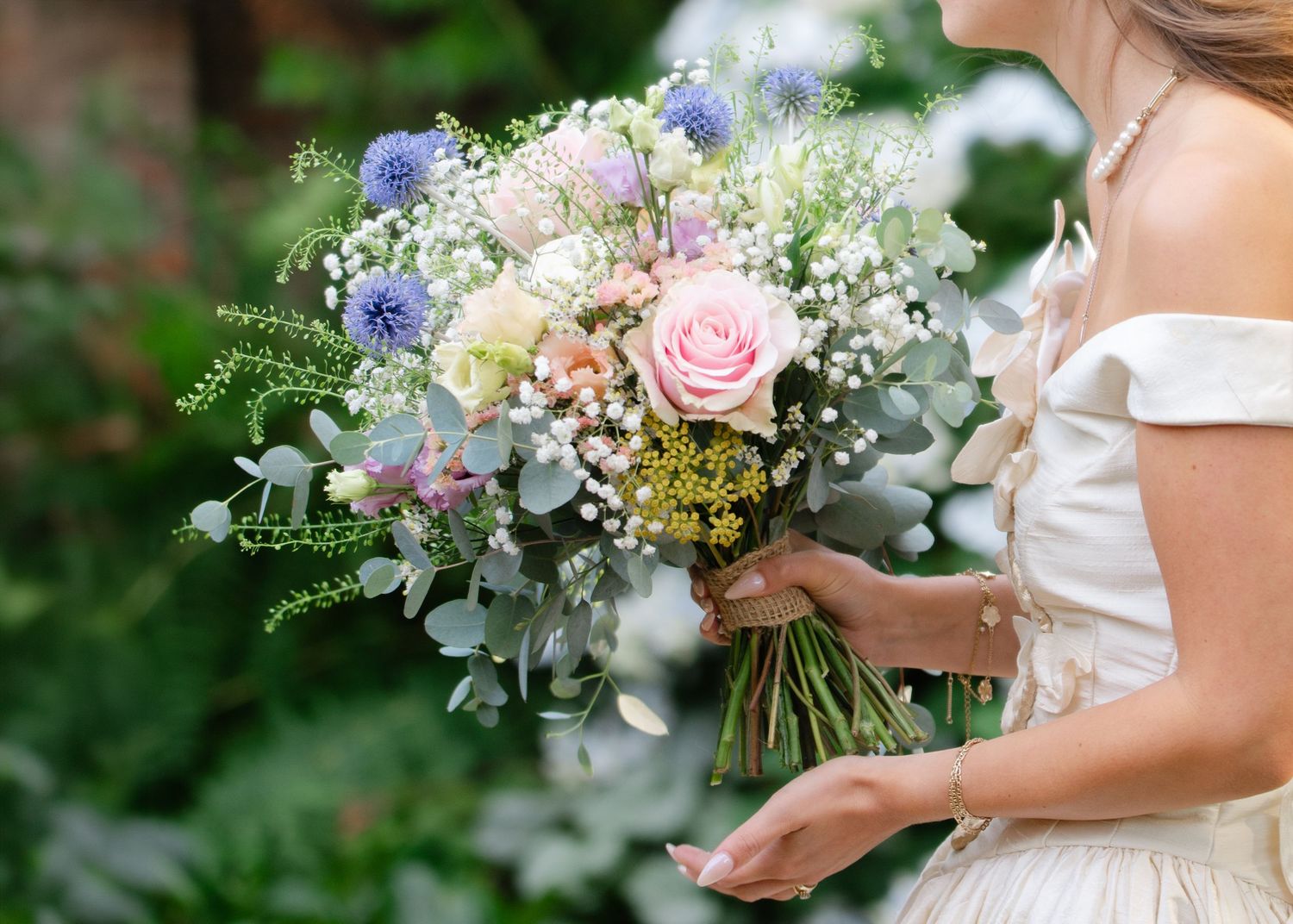 Bride holding a romantic wildflower wedding bouquet with pink roses, blue thistles, and eucalyptus in soft pastel tones.