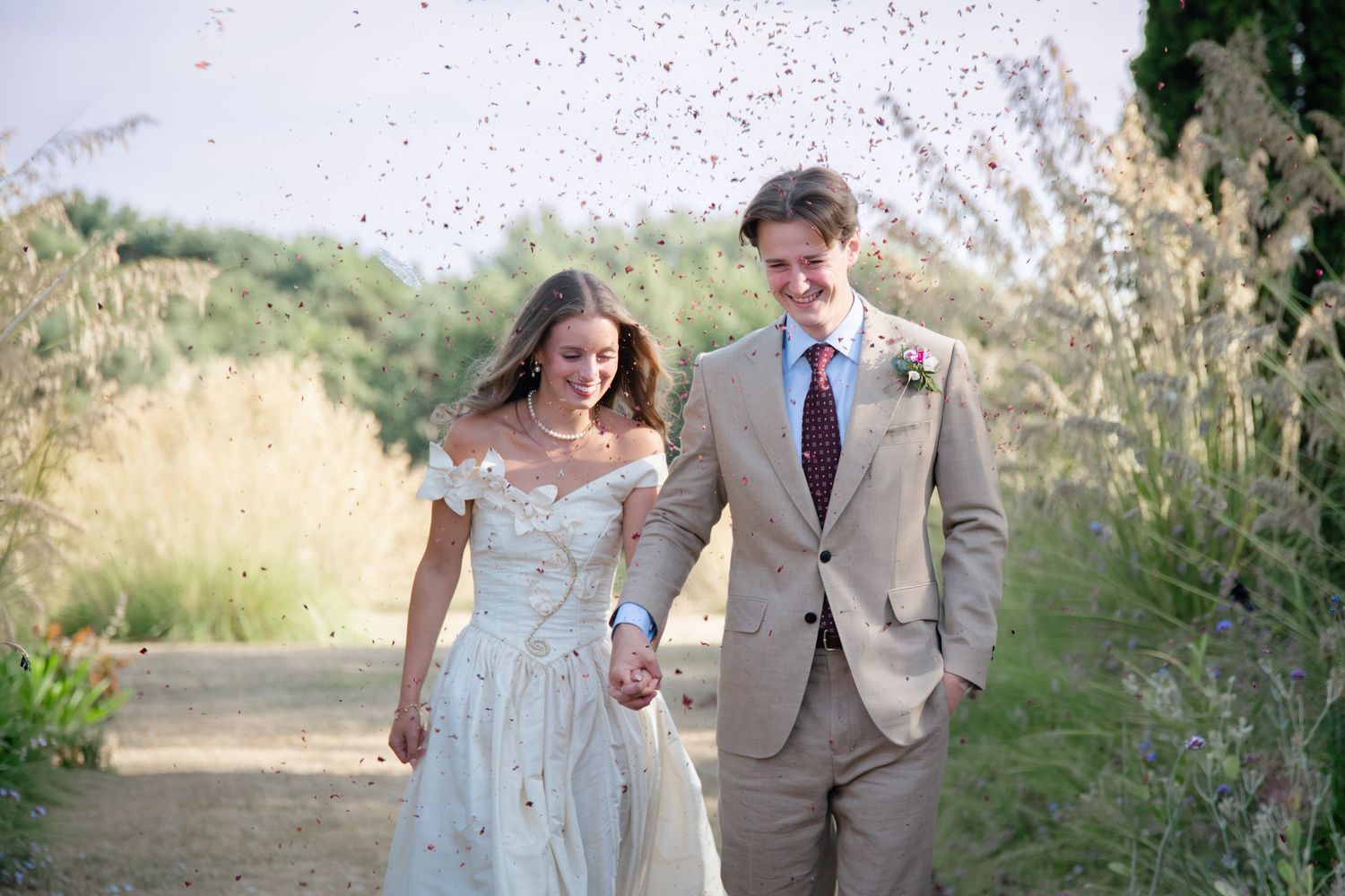 Newlywed couple walking hand in hand outdoors, bride in white off-shoulder gown, groom in tan suit, confetti falling.